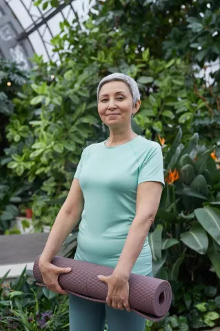Smiling senior woman with yoga mat in a vibrant greenhouse setting, promoting well-being.