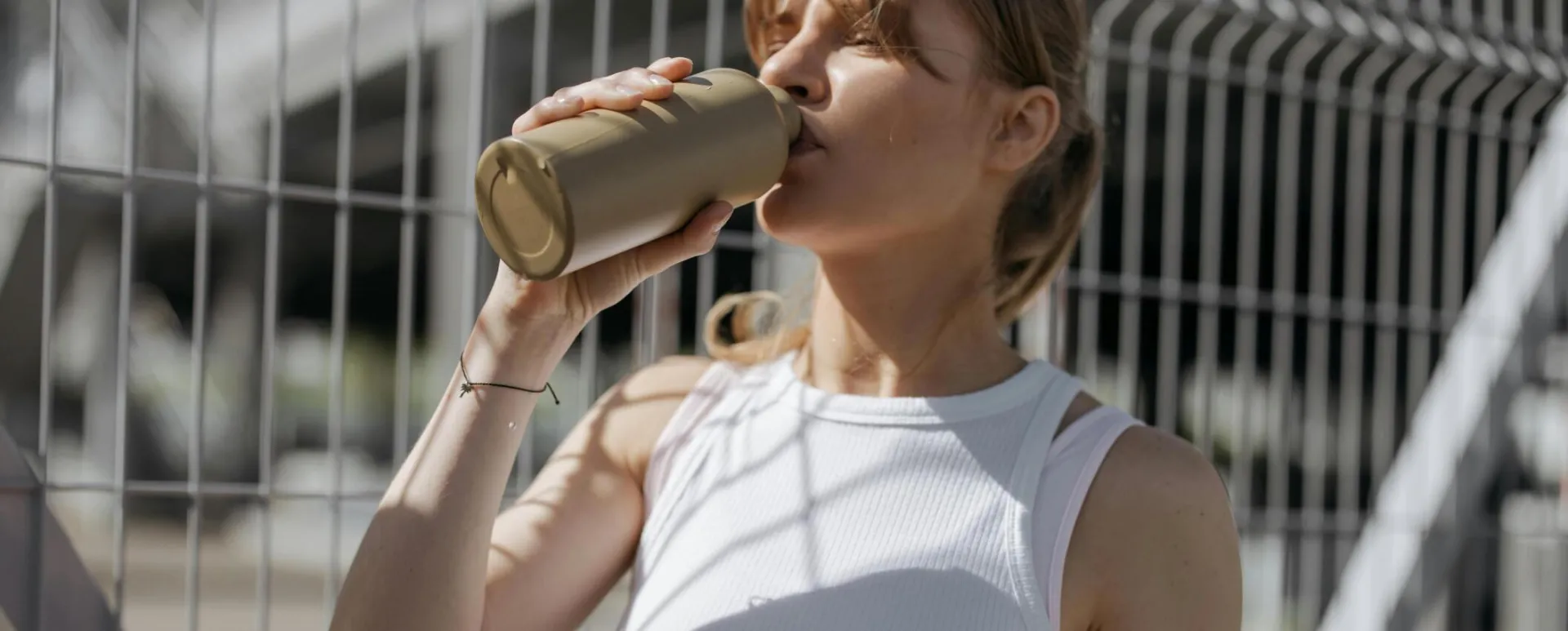 Woman in white tank top drinking from a tumbler outdoors on a sunny day.