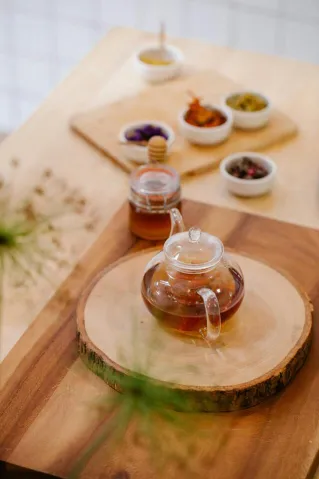 A glass teapot on a round wooden board surrounded by various tea ingredients in small bowls.