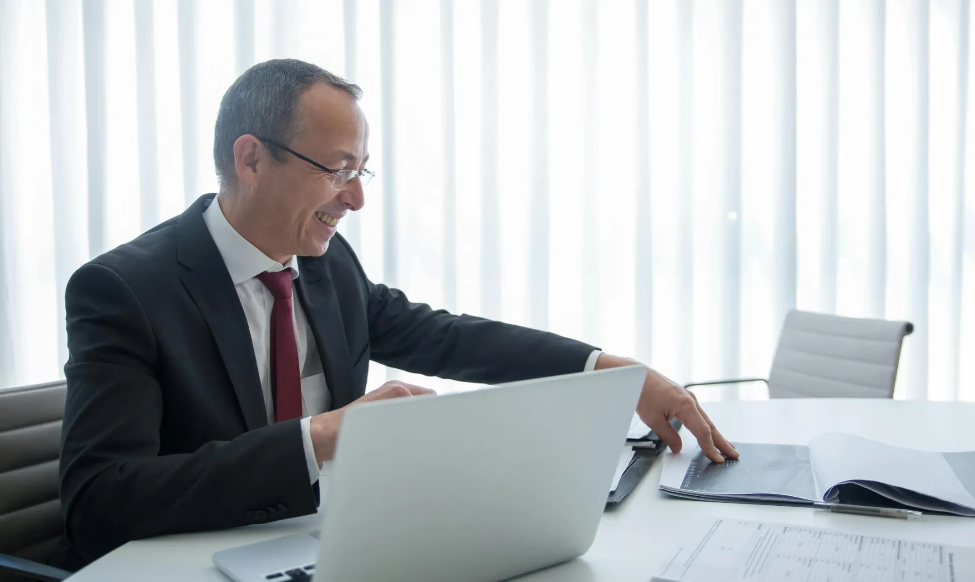 Elderly businessman in a suit working on a laptop and reviewing documents in a bright office setting.