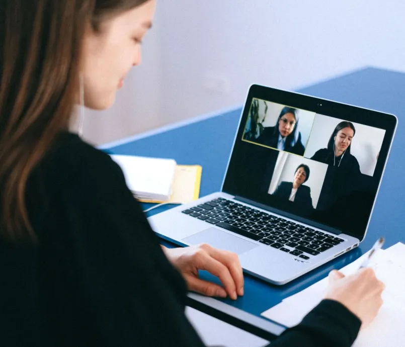 A woman engaging in a video conference using a laptop at home, taking notes.