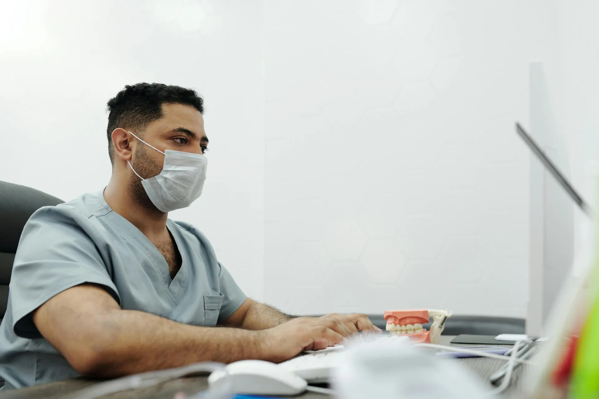 Medical professional in scrubs and mask working on computer in clinic office.