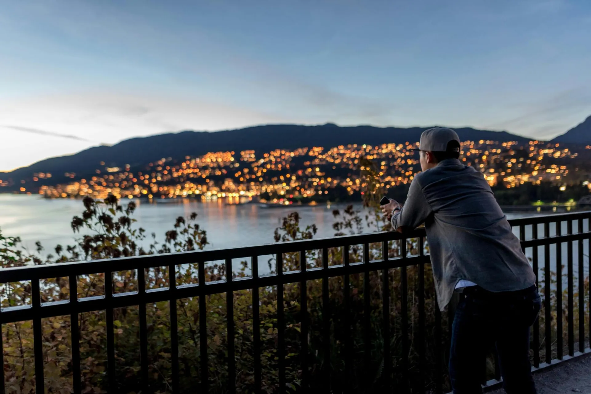 Adult man gazing at illuminated cityscape by the bay at dusk, capturing tranquility and scenic beauty.