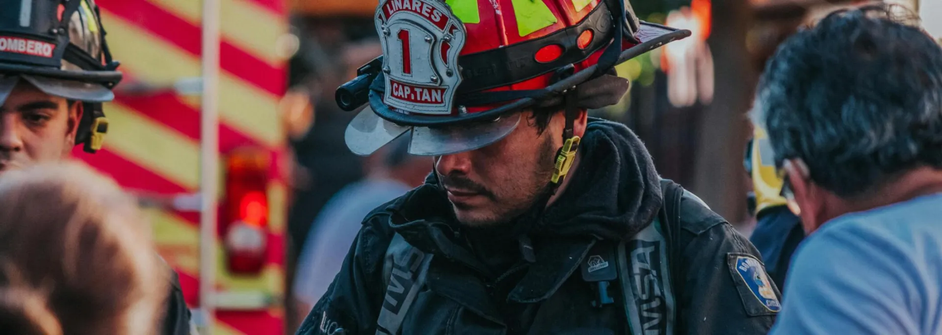 Firefighter attending an emergency on city streets, wearing uniform and helmet, with emergency vehicles in background.