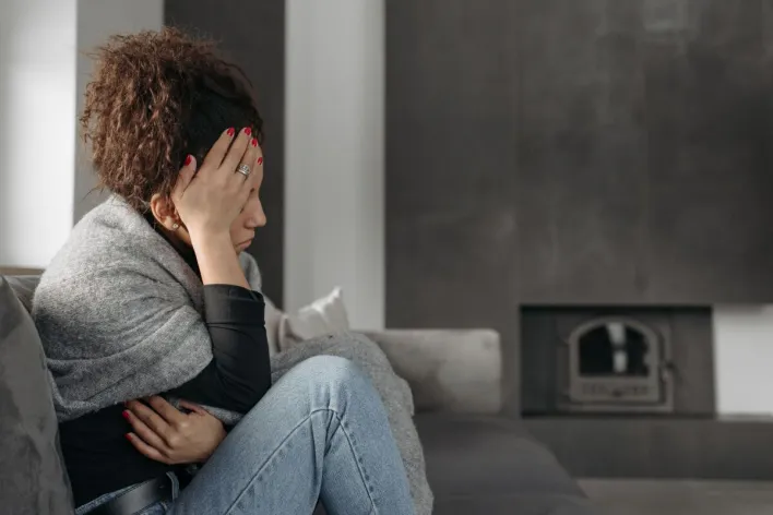 A woman sits on a couch indoors, wrapped in a blanket, holding her head in discomfort.