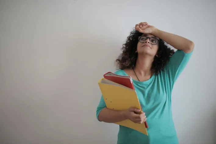 A woman in a teal top looks overwhelmed while holding colorful folders, symbolizing workplace stress.