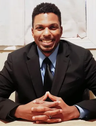 A professional black man smiling while seated by a window, embodying modern business vibes.
