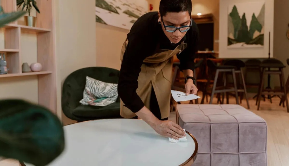 A barista wipes down a table in a stylish café interior, showcasing cleanliness and hospitality.