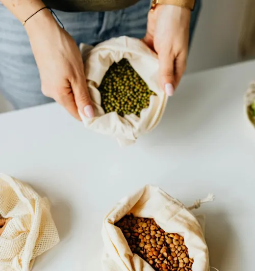 Woman holding reusable fabric bags filled with assorted beans on a white tabletop, promoting sustainability.