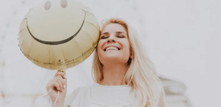 Cheerful woman holding a smiley balloon outdoors on a sunny day, exuding happiness and positivity.