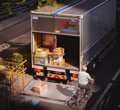 A delivery truck unloading boxes in a city street, with a bicyclist passing by on a sunny day.