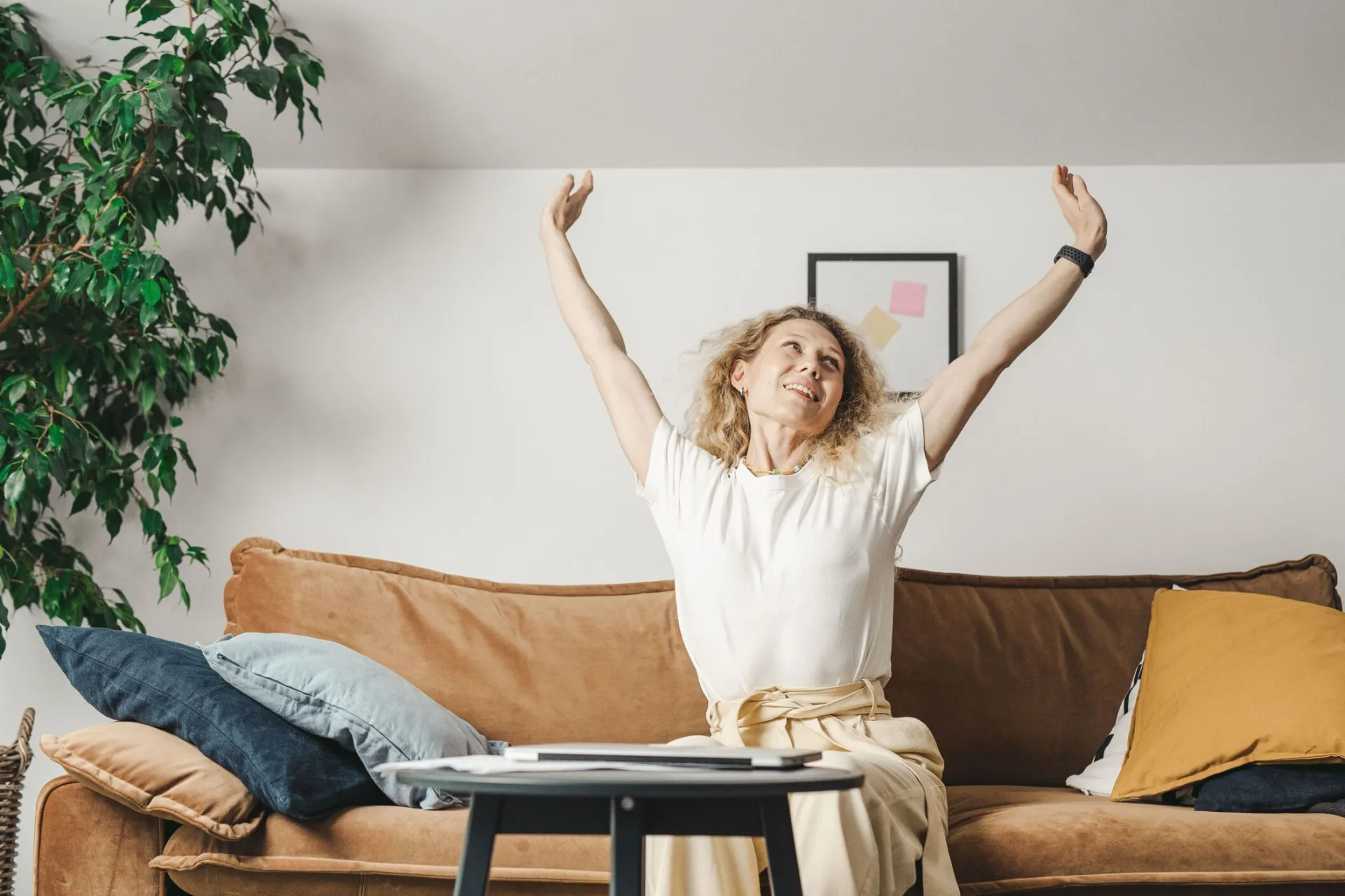 Smiling woman with curly hair raises her arms in joy while sitting on a cozy couch indoors.