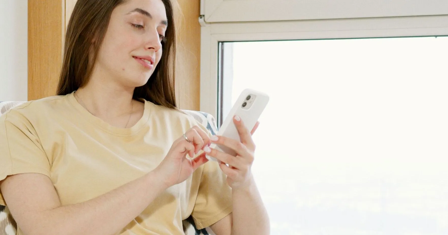 A woman sitting in a cozy armchair using her smartphone with a cup of coffee nearby.