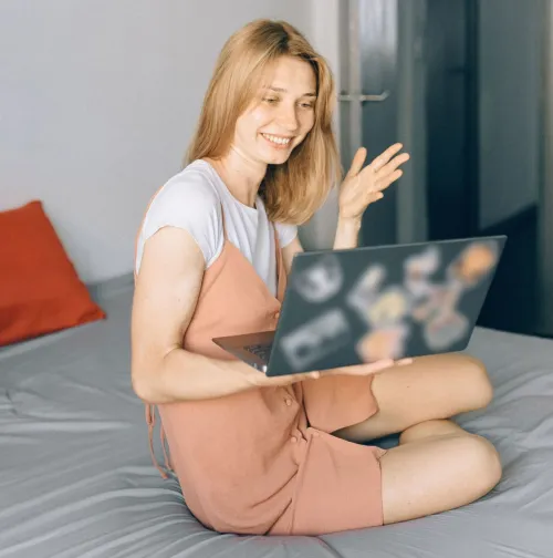 Woman sitting on bed working on laptop, smiling during a video call in cozy home setting.