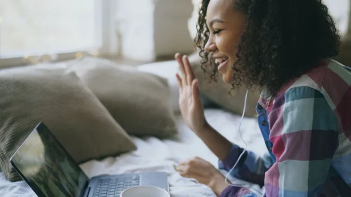 Cheerful woman waving during a video call from home laptop setup.