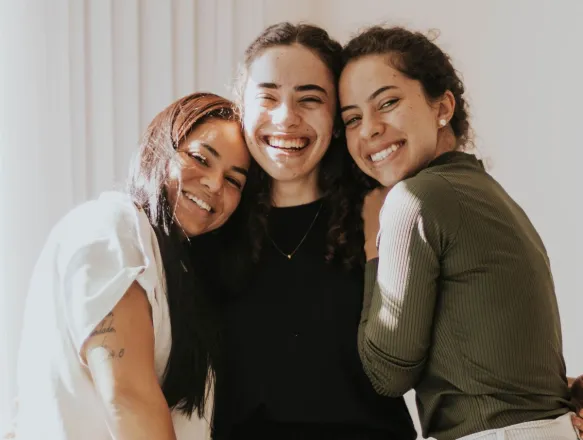 Three smiling friends hugging indoors, creating a warm and joyful atmosphere.