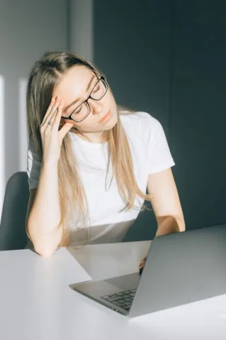 Young woman in glasses feeling tired while working on a laptop in an office setting.