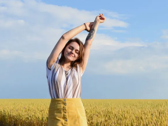 A young woman enjoying a sunny day, stretching her arms in a wheat field with a clear blue sky.