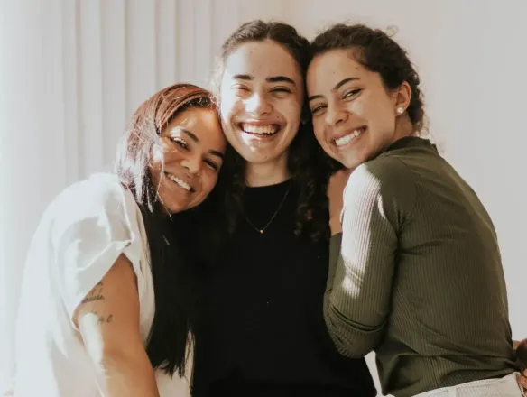 Three smiling friends hugging indoors, creating a warm and joyful atmosphere.