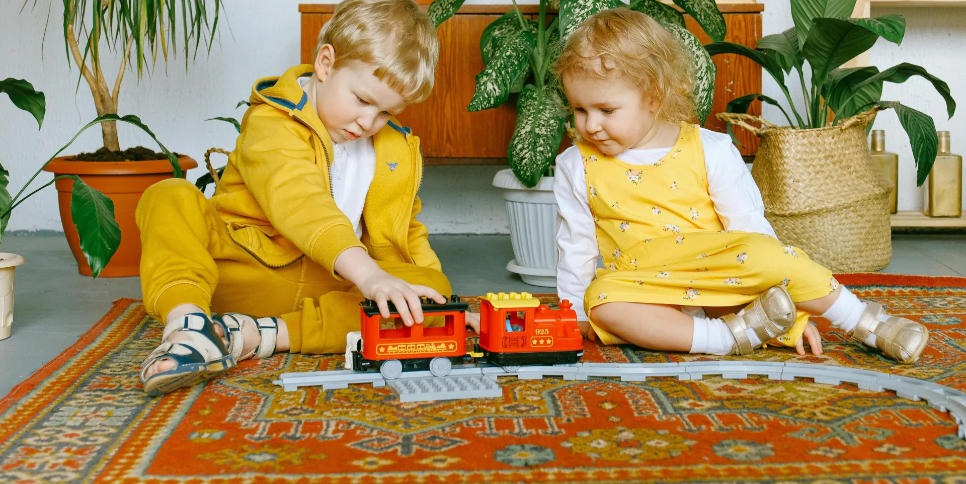Two children enjoy playing with a toy train on a colorful rug indoors surrounded by plants.