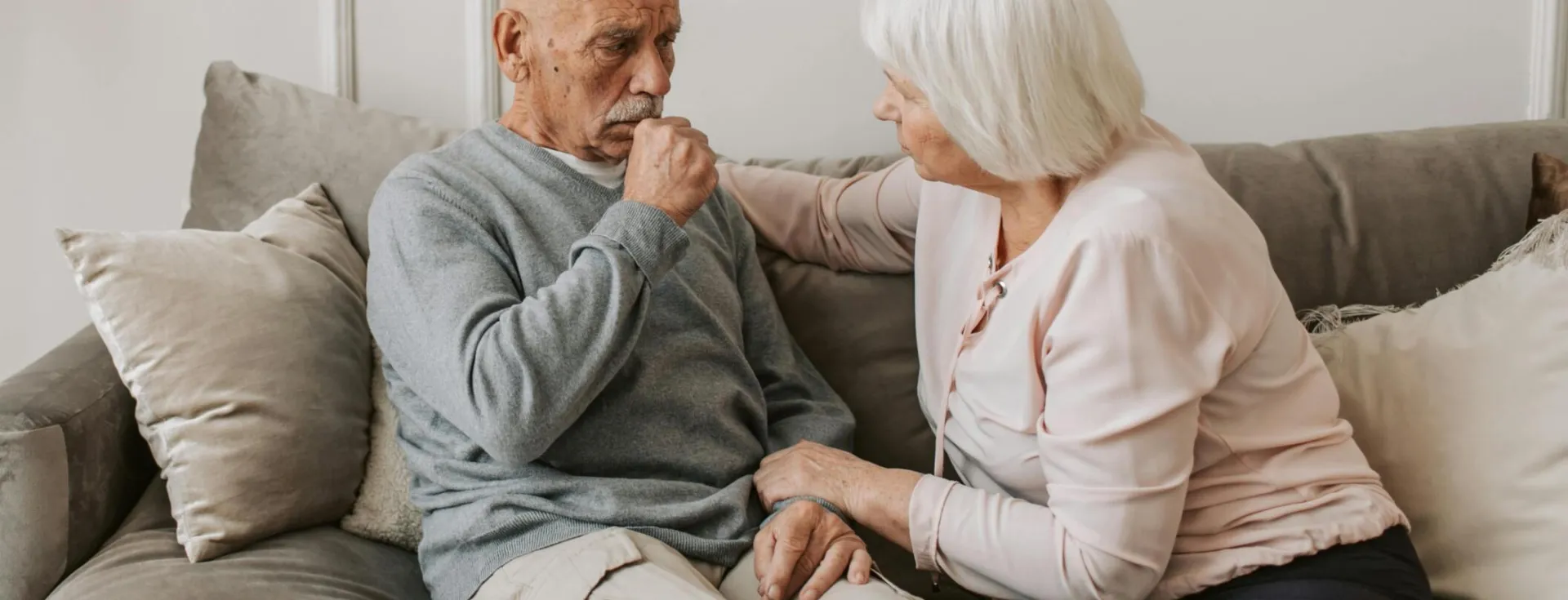 An elderly woman comforts a man coughing on a couch, showcasing care and affection.