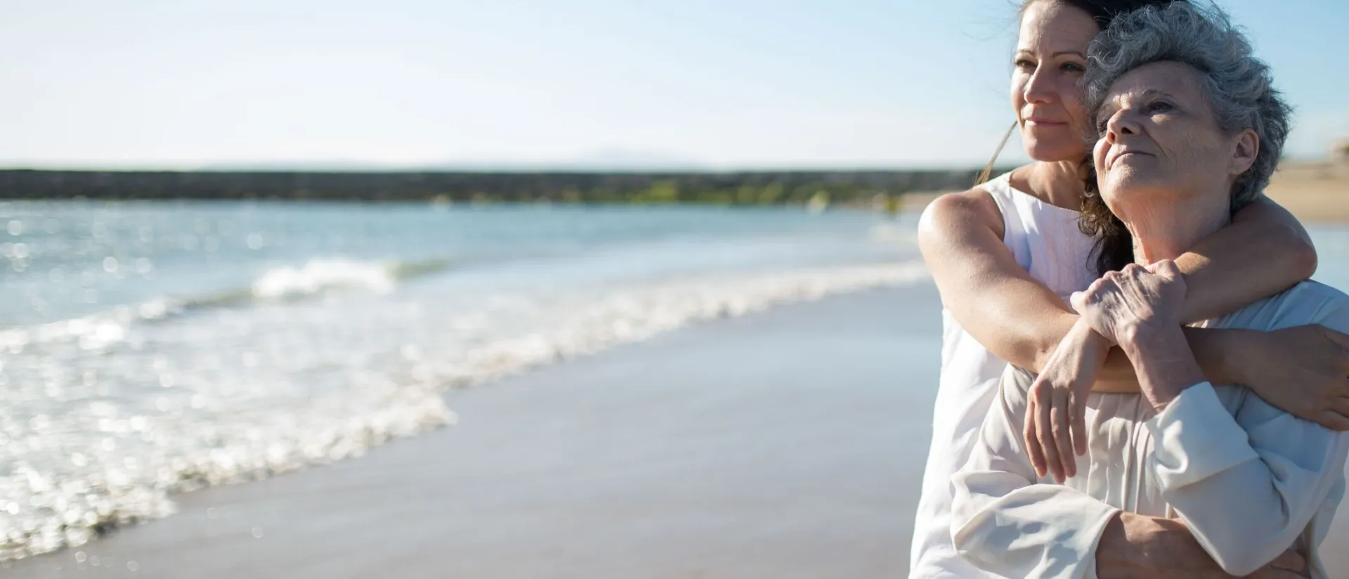 An elderly mother and adult daughter share a warm embrace on a sunny beach in Portugal.
