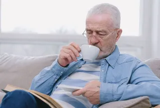 Elderly man enjoying a peaceful moment on sofa with a hot drink and book.