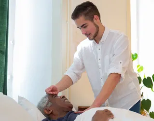 A young man supports an elderly man in bed, offering care in a bright room.