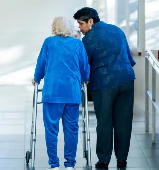 A nurse assists an elderly woman with a walker in a bright hospital corridor.