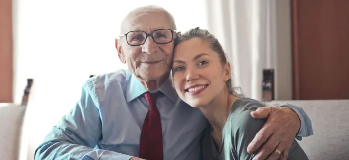 Positive senior man in formal shirt and eyeglasses hugging smiling young lady while sitting at table and looking at camera