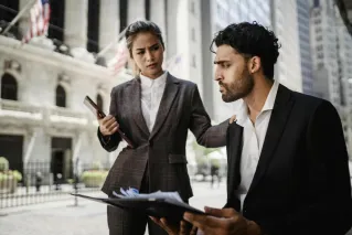 Two business professionals in formal attire discussing documents in a financial district setting outdoors.