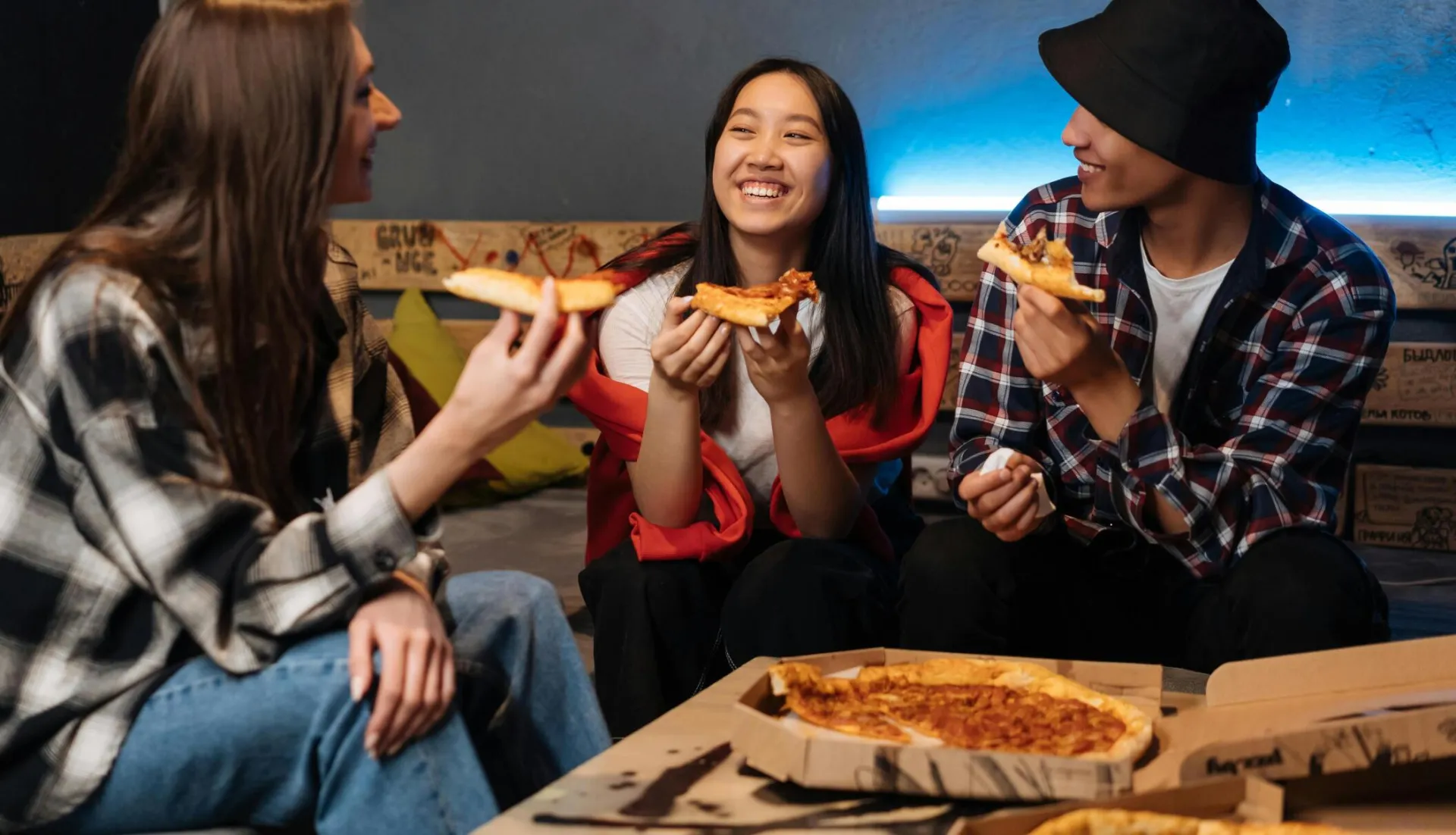 Three teenagers laughing and enjoying pizza together indoors, creating a joyful and friendly atmosphere.