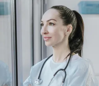 A female medical professional in a disposable gown looks thoughtfully out a window, stethoscope around her neck.
