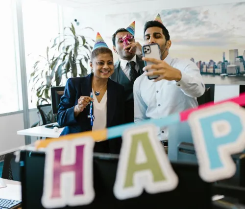 Three colleagues celebrating a birthday in the office with party hats and decorations, capturing a fun selfie.