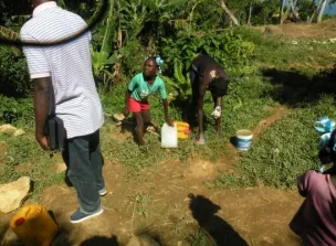 A young girl with braided hair carries a jug of water on her head in a rural area of Haiti.