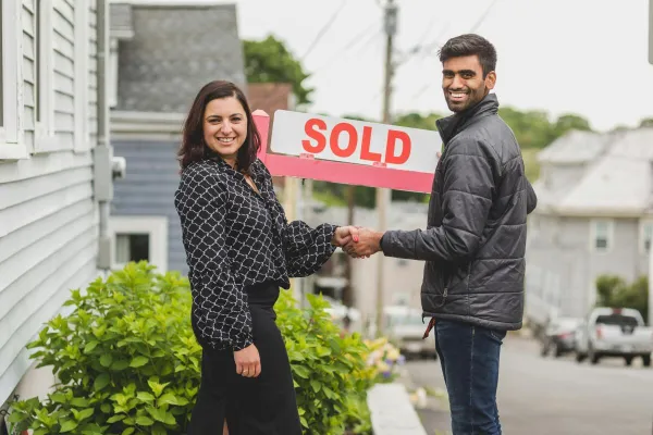 Two people shake hands in front of a sold sign, celebrating a real estate deal.