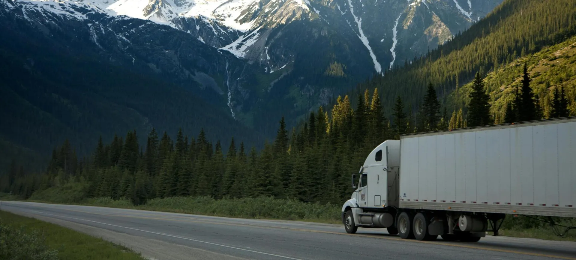 A semi-truck travels along a highway with snow-capped mountains in the background.