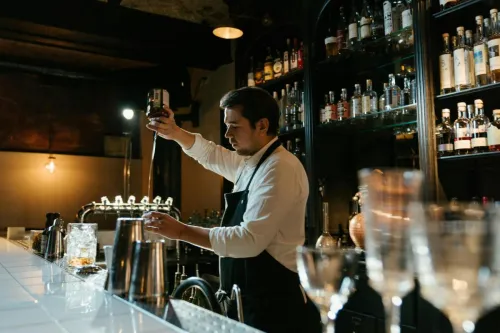 A bartender expertly pouring a drink at a bar, surrounded by various bottles and tools.