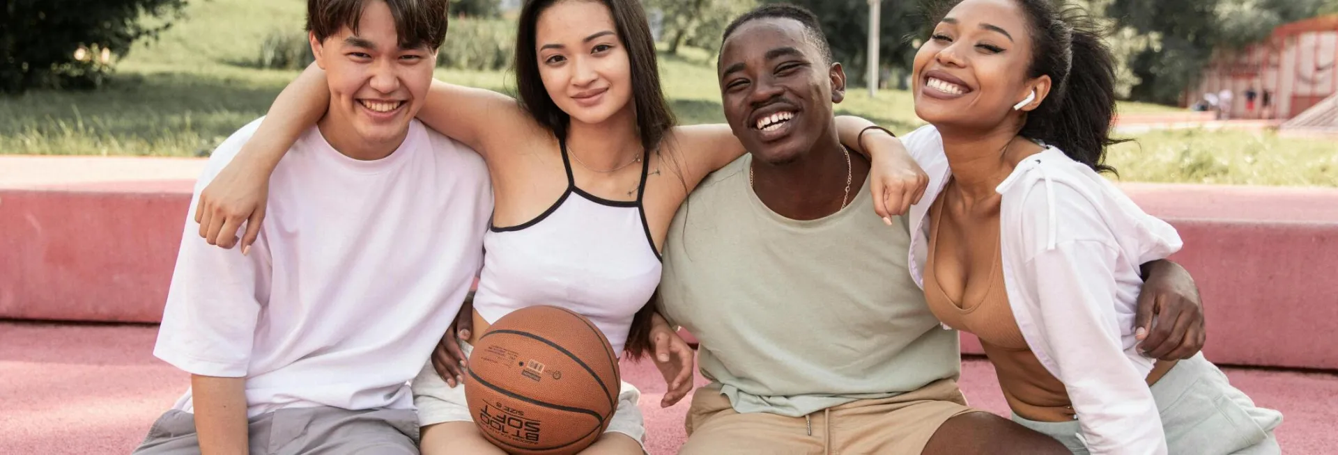 Content young multiracial friends wearing casual clothes embracing and looking at camera with happy smiles while sitting on bench in green park