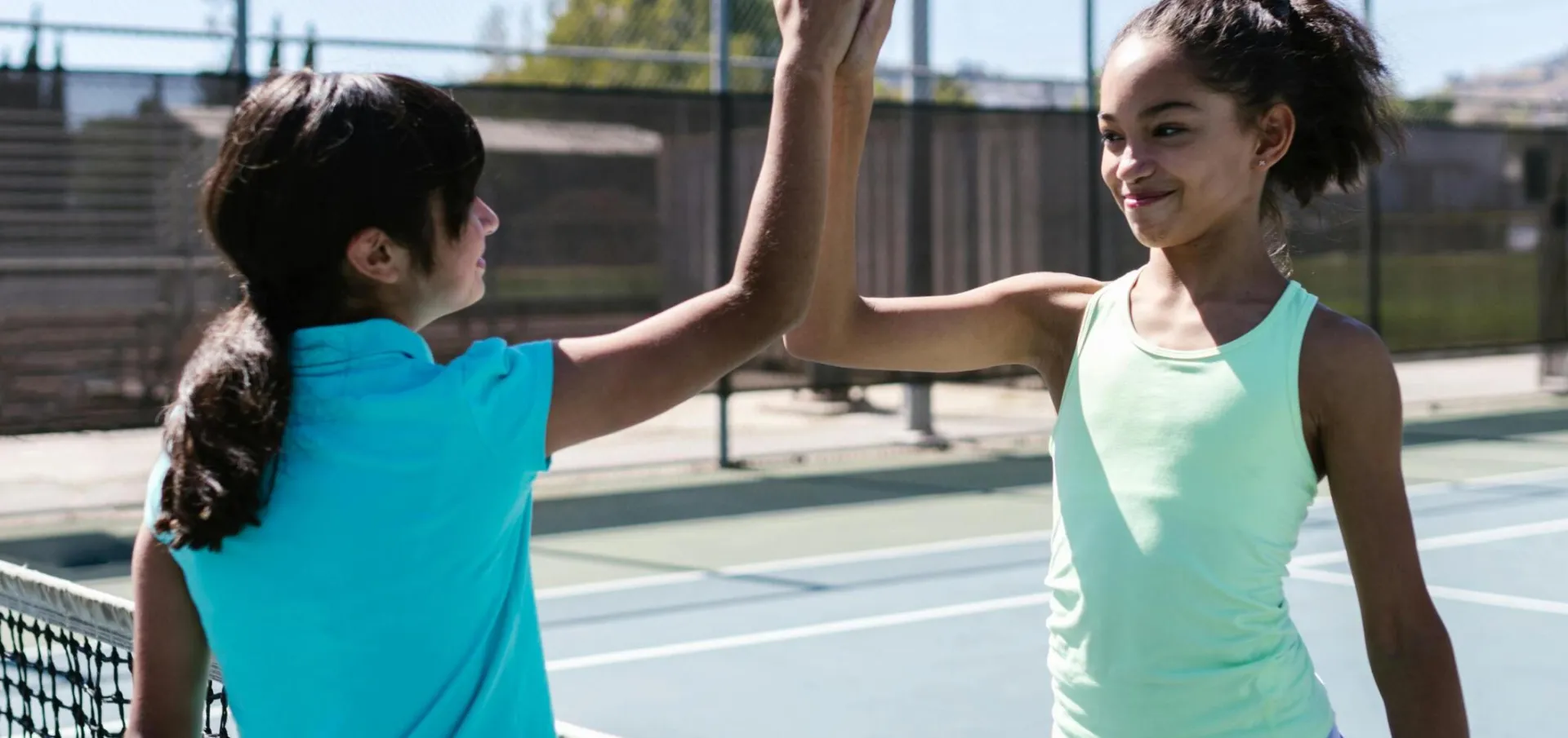 Two teen girls celebrating with a high five on a sunny tennis court.