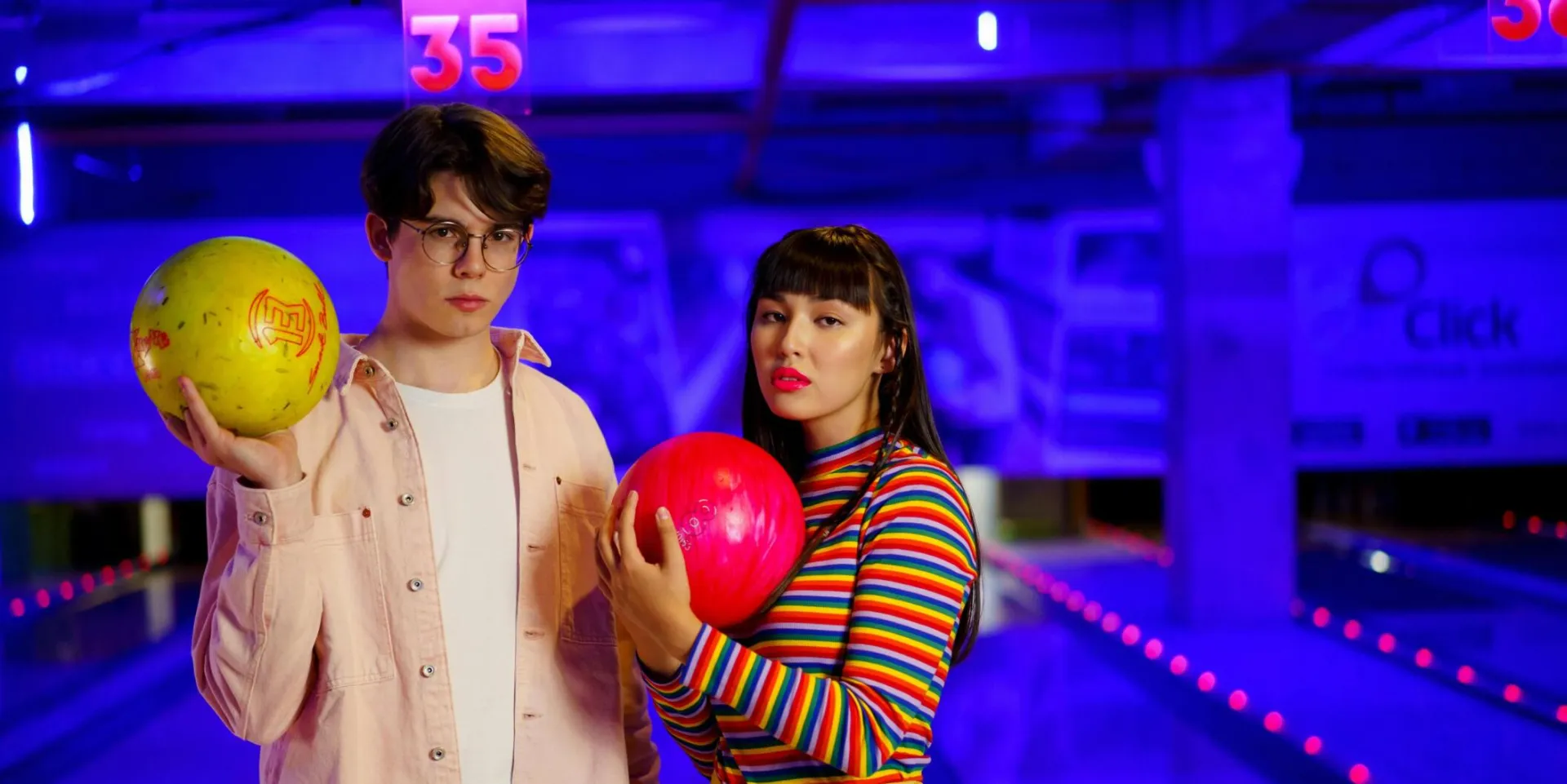 Two stylish young adults holding bowling balls at an illuminated alley.
