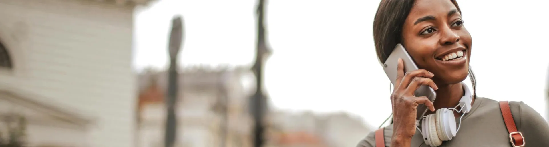 Young woman with headphones smiling while talking on phone outdoors in city.