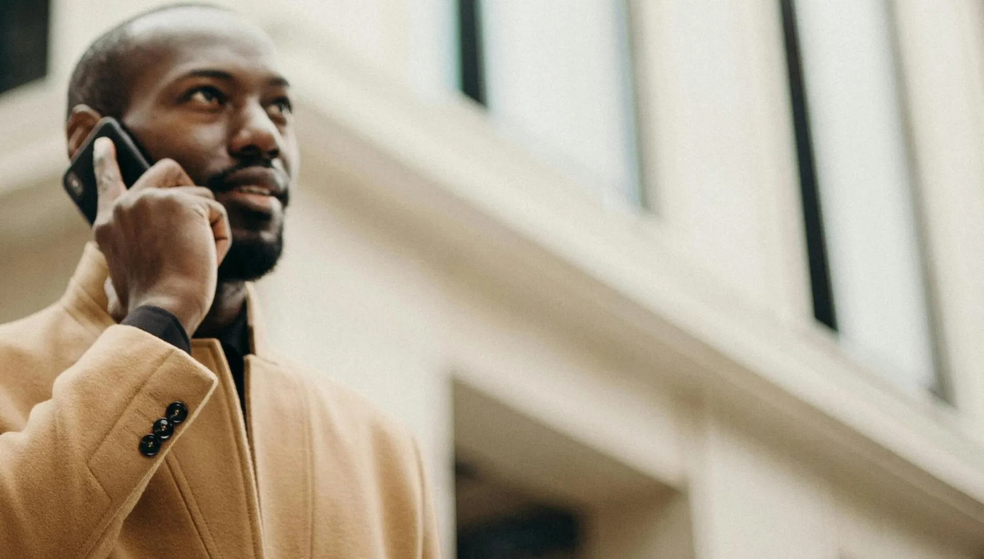 Confident man in a trench coat making a call outdoors in front of a modern building.