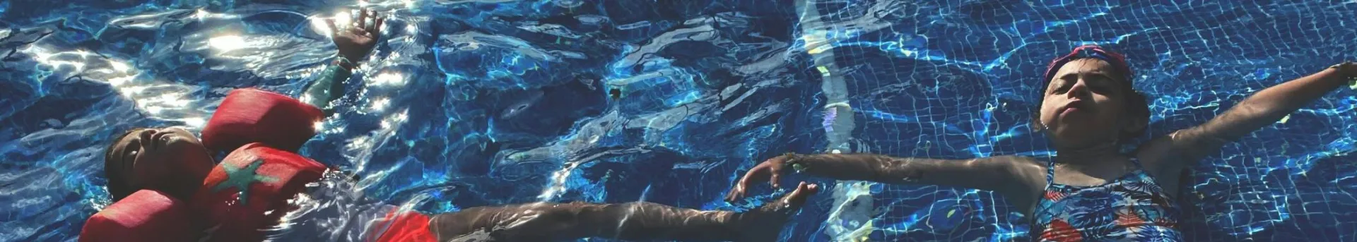 Two children relaxing and floating in a swimming pool with water wings under bright sunlight.