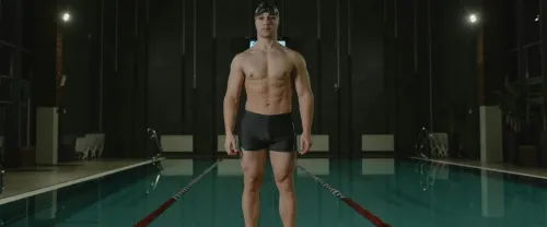 Athletic male swimmer standing confidently poolside indoors.