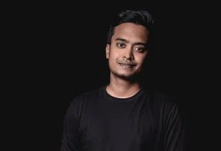 Studio portrait of a young South Asian man in a black shirt against a dark background.
