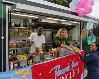 A man and woman stand in front of a food truck, offering street food at night.