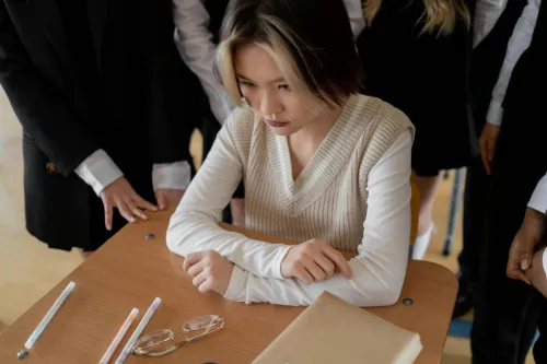 Teen girl looks thoughtful while sitting alone at a school desk surrounded by classmates.