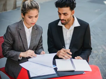 Two professionals in business suits reviewing documents while sitting outdoors at a red table.