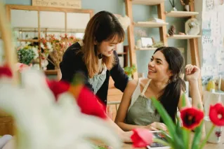 Happy young multiracial female florists wearing uniforms having conversation in light cozy floristry store and looking at each other with smiles while sitting at table with tablet